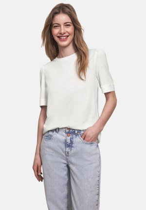 Junge Frau, die lächelt, trägt ein weißes kurzärmeliges Oberteil und hellblaue, hoch taillierte Jeans, mit einer Hand in der Tasche.