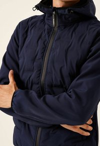 Man i en marinblå huvenjacka med blixtlås fram, med armarna korsade, mot en ljus bakgrund.