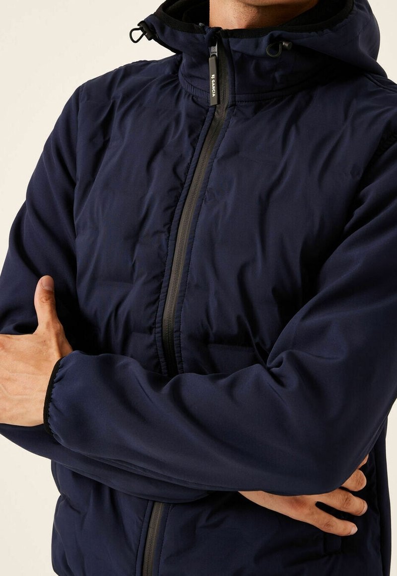 Man i en marinblå huvenjacka med blixtlås fram, med armarna korsade, mot en ljus bakgrund.