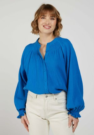 Femme souriante portant un chemisier bleu vif à manches longues boutonné avec des poignets froncés et un pantalon crème taille haute sur un fond clair.