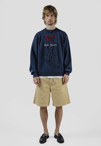 Laivastonsininen neulepaita, jossa on kirjailtu hiihtokuvio ja punainen korostus kauluksessa, yhdistettynä beigen sävyisiin shortseihin ja mustiin loafer-kenkiin. Yksinkertainen, rento asu.