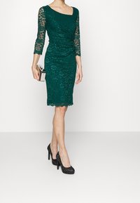 Robe en dentelle vert émeraude à manches trois-quarts, design plissé et ourlet festonné, associée à des talons hauts noirs pailletés et une pochette argentée.