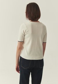 Pull à manches courtes blanc côtelé avec un col rond, un motif texturé et une bordure contrastante noire. Un jean bleu foncé avec des poches arrière complète la tenue.