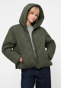 Giacca cappuccio trapuntata verde oliva, con chiusura con zip, maniche lunghe e tasche laterali, abbinata a una maglietta grigia e jeans.