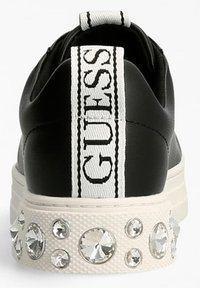 Sneaker nera con suola bianca texturizzata, dotata di un'etichetta a strappo bianca con il logo e di una fila decorativa di strass trasparenti attorno alla base.