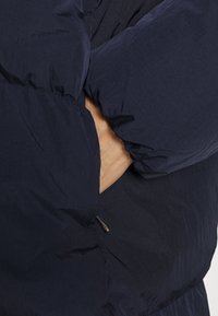 Tmavě modrá péřová bunda s texturovaným povrchem, vybavená vysokým stojáčkem a kapsami na zip. Rukávy jsou nařasené pro větší teplo.