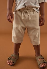 Smėlinės spalvos medvilninės šortai su elastine liemene ir reguliuojamu raišteliu. Derinami su rudos spalvos sandalais, kuriuose yra dvi sagtelės šonuose.