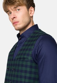 Zielona i granatowa kamizelka w kratę o podwójnym zapięciu, z klapkami owalnymi i subtelną teksturą, noszona na ciemnoniebieskiej koszuli.