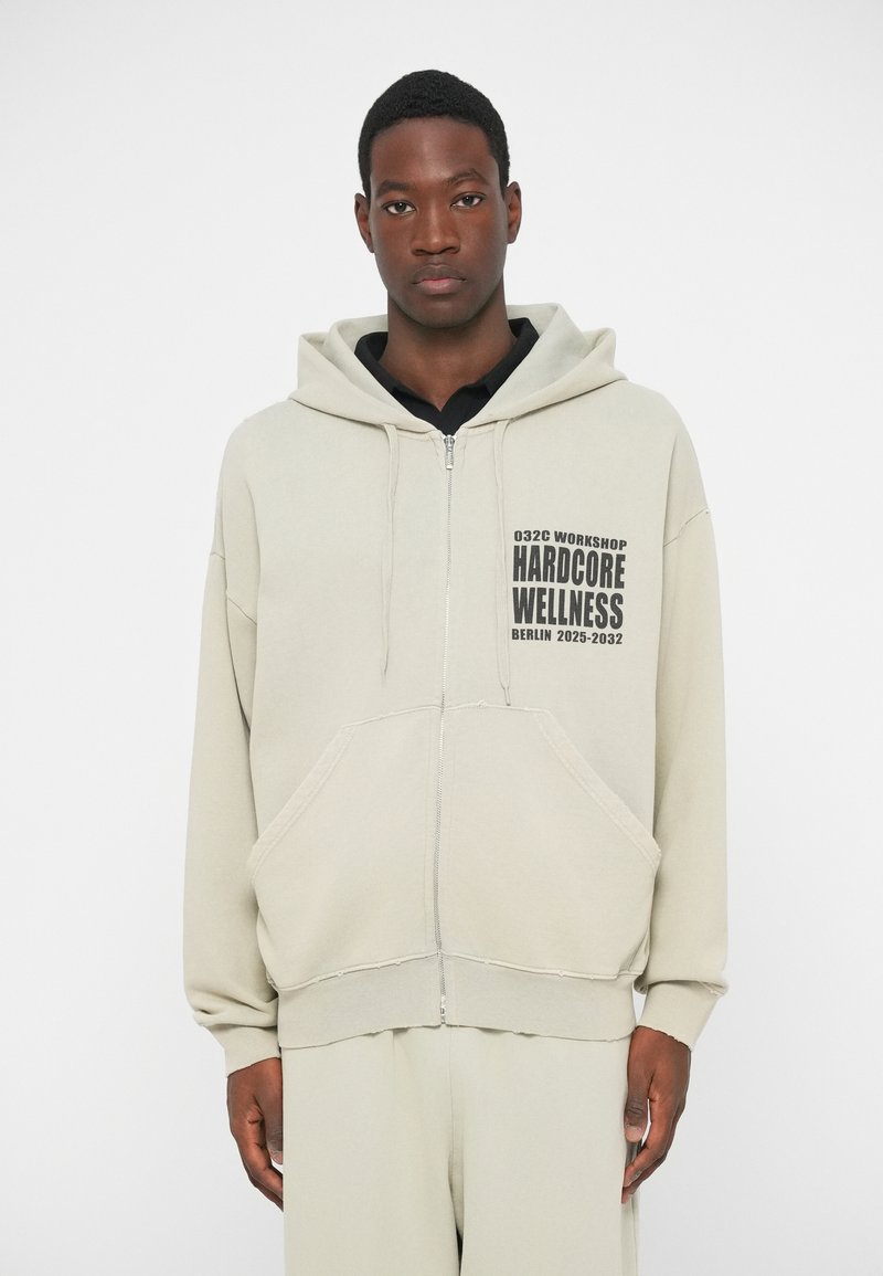 Lichtgrijze zip-up hoodie gemaakt van zachte stof. Heeft een voorzak, een capuchon met trekkoord en een geprinte logo aan de linkerkant.