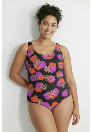 Mujer sonriente con cabello rizado que lleva un traje de baño de una pieza negro con patrones abstractos en naranja, púrpura y estampado de leopardo, de pie frente a una pared blanca.