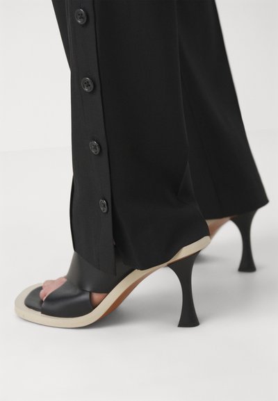 Black high-heeled sandals with a peep-toe design, featuring a smooth black upper and a contrasting beige sole. Buttons from black trousers are visible.