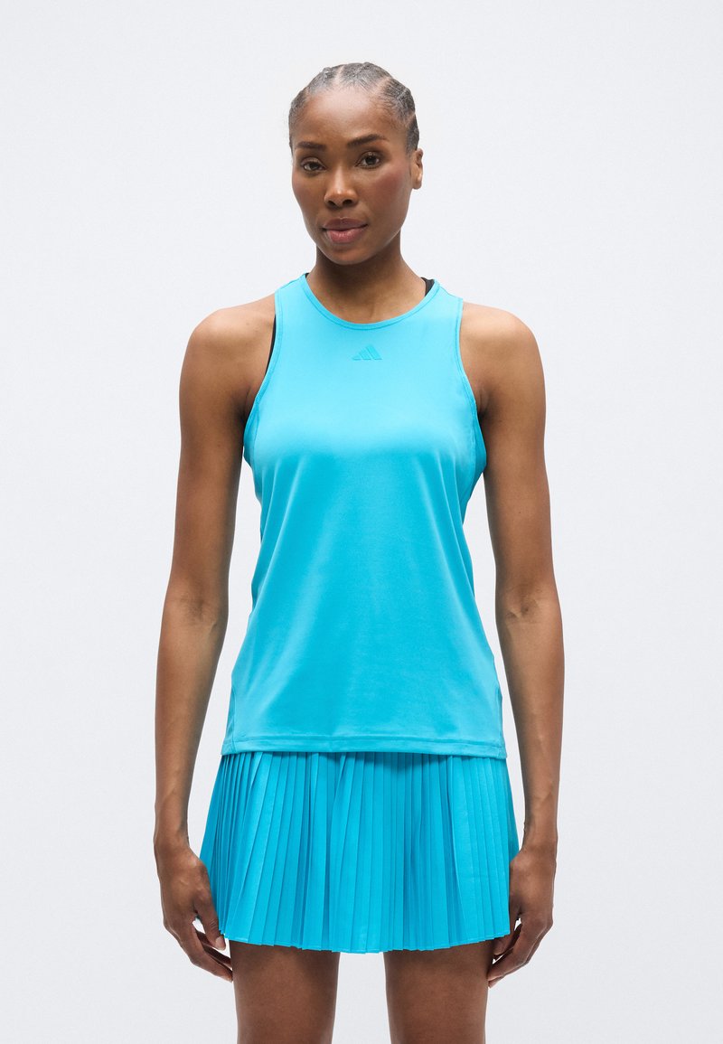 Ένα τυρκουάζ αθλητικό τοπ χωρίς μανίκια σε συνδυασμό με μια πλισέ φούστα, κατασκευασμένα από ελαφρύ ύφασμα, με στρογγυλή λαιμόκοψη και διακριτικό branding.