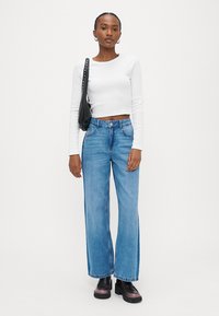 Γυναίκα που φοράει λευκή μακρυμάνικη κοντή μπλούζα, τζιν παντελόνι υψηλής μέσης με φαρδιά πόδια μπλε, μαύρα παπούτσια πλατφόρμα και κρατάει μαύρη τσάντα ώμου.