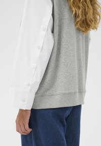 Sweat-shirt gris avec manches de chemise blanches boutonnées. Texture douce, coupe décontractée et ourlet côtelé. Tissu bleu visible à la taille.