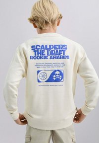 Sudadera color crema con texto azul llamativo en la parte trasera, gráfico de baloncesto e ícono de calavera; hecha de tela suave y texturizada.