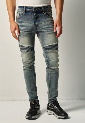 Hombre que lleva vaqueros pitillo azules desteñidos con parches acanalados en las rodillas y zapatos deportivos negros, de pie contra un fondo liso.