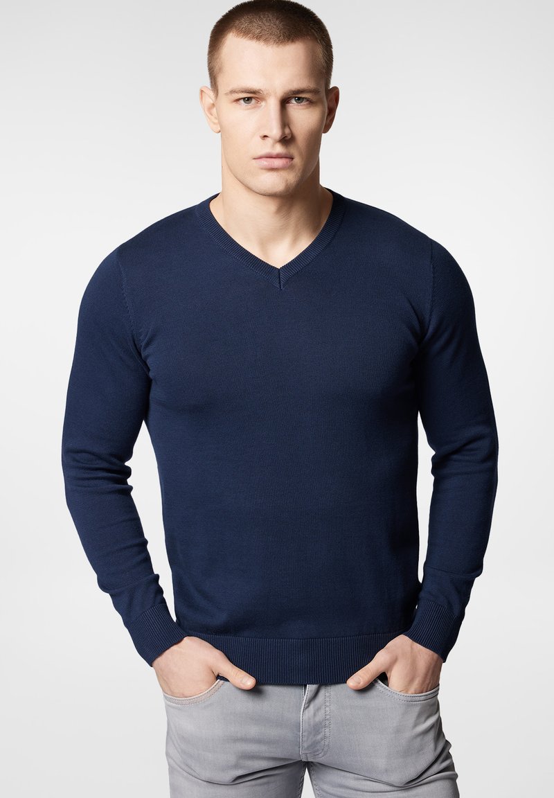 Granatowy sweter w serek wykonany z lekkiej dzianiny, z długimi rękawami oraz ściągaczami przy rękawach i u dołu, noszony na jasnoszare spodnie.