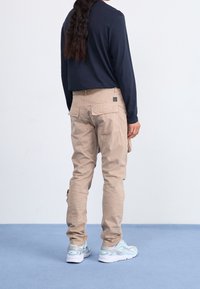 Osoba z długimi ciemnymi włosami nosi beżowe spodnie cargo, granatową koszulę z długim rękawem oraz jasnoniebieskie i białe sneakersy, stojąc na niebieskiej podłodze.