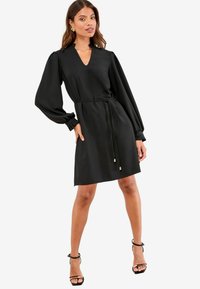 Schwarzes Kleid mit V-Ausschnitt, langen Puffärmeln und einem Taillengürtel. Hergestellt aus glattem Stoff mit lässiger Passform, verziert mit goldenen Akzenten an den Enden des Gürtels.
