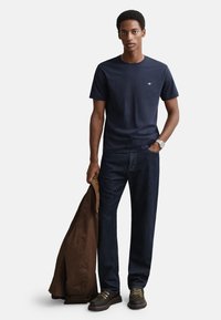 Marinblå t-shirt i bomull med rund hals, kombinerad med mörka jeans. Brun jacka hålls i vänster hand, bär snörkängor. Enkel design, avslappnad passform.