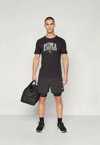 Black sports t-shirt with "PUMA sports club" graphic, paired with black shorts and a black duffel bag. Footwear features black accents.