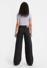 Svarta vida jeans av denim, med en framträdande bakficka, tillsammans med en figursydd kortärmad mönstrad topp. Enfärgad bakgrund.