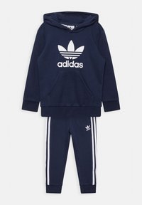 Set di felpa e pantaloni della tuta blu navy, realizzato in tessuto morbido. La felpa presenta una tasca frontale e un grande logo Adidas bianco; i pantaloni hanno strisce bianche.