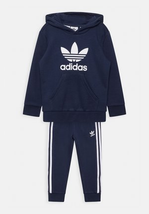 Set di felpa e pantaloni della tuta blu navy, realizzato in tessuto morbido. La felpa presenta una tasca frontale e un grande logo Adidas bianco; i pantaloni hanno strisce bianche.
