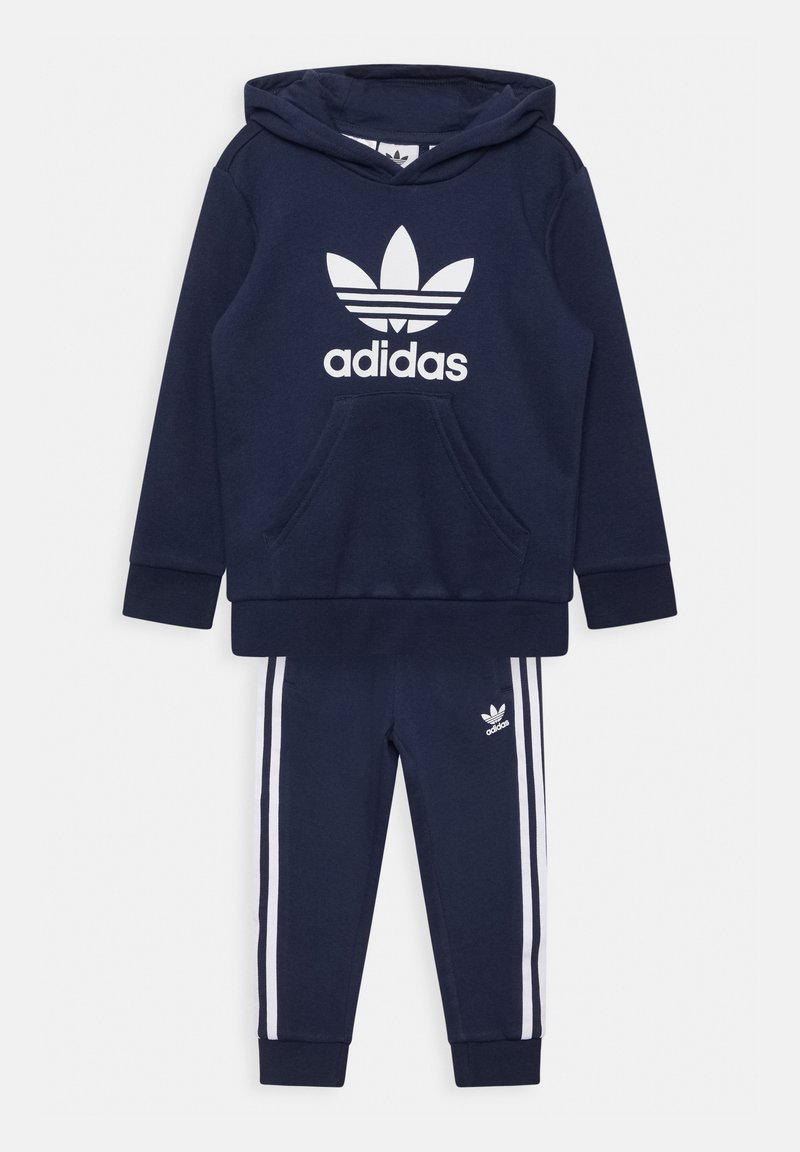 Set di felpa e pantaloni della tuta blu navy, realizzato in tessuto morbido. La felpa presenta una tasca frontale e un grande logo Adidas bianco; i pantaloni hanno strisce bianche.