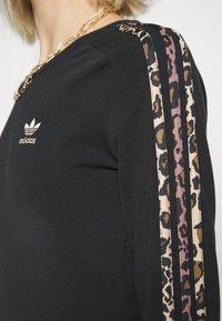 Černá mikina s kulatým výstřihem, zlatým logem Adidas na hrudi a pruhy s leopardím vzorem podél ramen.