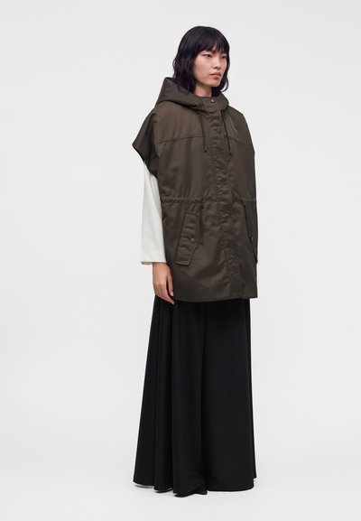 Personne portant un gilet à capuche vert foncé avec de grandes poches par-dessus un haut blanc à manches longues et un pantalon noir large, debout devant un fond uni.