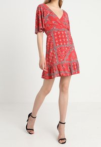 Rotes kurzes Kleid mit V-Ausschnitt, Puffärmeln und einem gemusterten Design, das Paisleymuster und geometrische Formen zeigt. Rüschenbesatz am Saum, glattes Material.