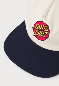 Beige sapka sötétkék karimával. Elöl hímezett piros és sárga "Santa Cruz" logóval rendelkezik. Strukturált kialakítás, pamut anyag.