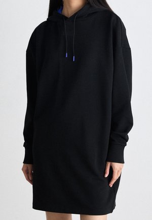 Czarna, oversize sukienka-bluza z kapturem, z troczkami zakończonymi niebieskimi końcówkami, długimi rękawami i ściągaczami przy mankietach, wykonana z miękkiej bawełny.