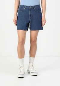 Denim shorts, blue color, high-waisted design, front pockets, raw hem, worn with white sneakers and socks, standing against a plain background.