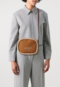 Hnědá kožená crossbody taška s umělým kožešinovým lemem, obdélníkového tvaru a vyraženým logem "JOOP! JEANS". Nosit ji na sobě personě v kostkovaném oblečení.