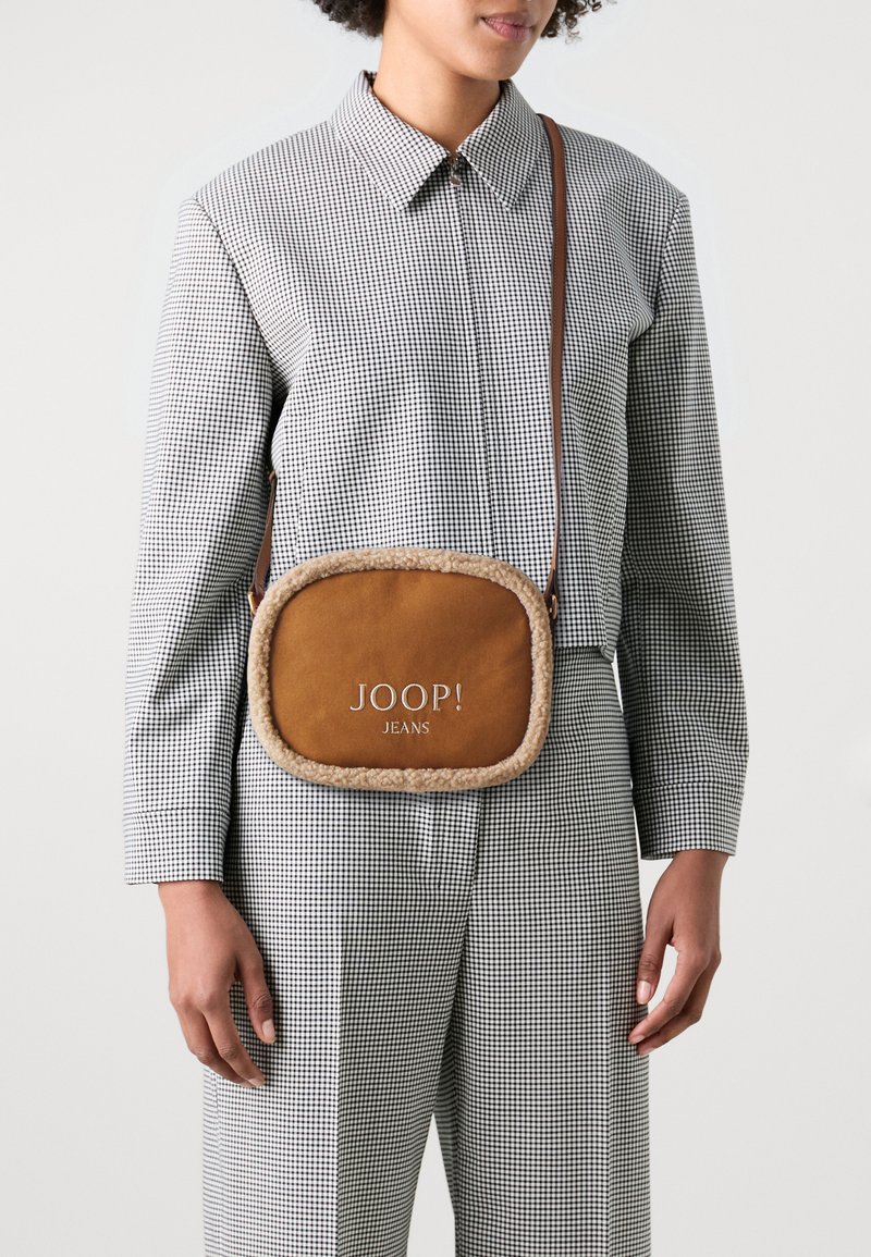 Hnědá kožená crossbody taška s umělým kožešinovým lemem, obdélníkového tvaru a vyraženým logem "JOOP! JEANS". Nosit ji na sobě personě v kostkovaném oblečení.