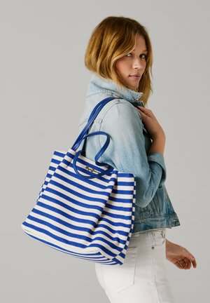 Femme en veste en jean claire et pantalon blanc tenant un sac cabas à rayures bleues et blanches sur l'épaule, devant un fond gris.