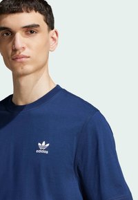 Tamsiai mėlynas medvilninis marškinėlis su apvaliu iškirpimu, trumpomis rankovėmis ir baltu Adidas logotipu kairėje krūtinės pusėje. Lygus paviršius.