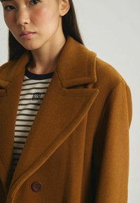 Manteau en laine marron avec un large col et une fermeture à double boutonnage, porté par-dessus une chemise rayée présentant un mélange de lignes horizontales.