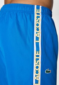 Blauwe sportshorts van meshmateriaal, met een witte streep met "LACOSTE" in het geel en een groen krokodillenlogo aan de zijkant.