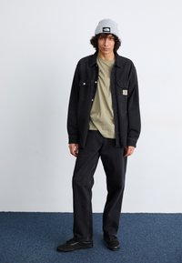 Camicia di denim nera con due tasche sul petto sopra una maglietta verde chiaro, abbinata a jeans neri e scarpe nere stringate. Berretto di maglia grigio.
