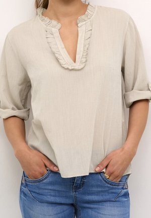 Hellbeige Bluse mit einem Rüschenausschnitt und lockerer Passform. Mit umgeschlagenen Ärmeln. Kombiniert mit blauen Jeans. Glatte Textur.