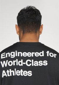 T-shirt de sport noir à manches courtes, avec un texte blanc audacieux au dos indiquant « Conçu pour les athlètes de classe mondiale ».