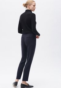 Maglione nero a collo alto abbinato a pantaloni taper navy. Design su misura con tasche posteriori e mocassini neri, su uno sfondo bianco minimale.