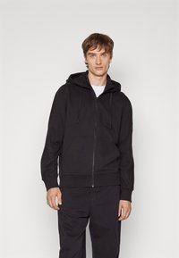 Sudadera con cremallera negra, capucha ajustable, mangas largas, puños acanalados y un bolsillo frontal. Textura suave, diseño minimalista.