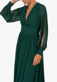 Grünes V-Ausschnitt-Kleid mit langen, transparenten Ärmeln und taillierter Sanduhrsilhouette. Verfügt über einen gerafften Rock und Knopfdetails an den Manschetten. Glatte Textur.