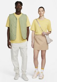Vesta matlasată verde, tricou galben, pantaloni cargo albi. Femeie în tricou galben, fustă bej, geantă transversală, adidași cu model, șosete albe.