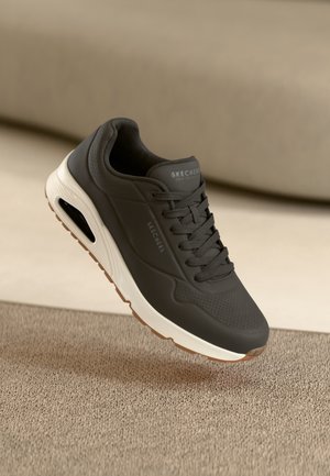 Tummanharmaa Skechers-tossu, jossa valkoinen pehmustettu pohja ja näkyvä ilma-aukko, esillä beigeä kangasta vasten.