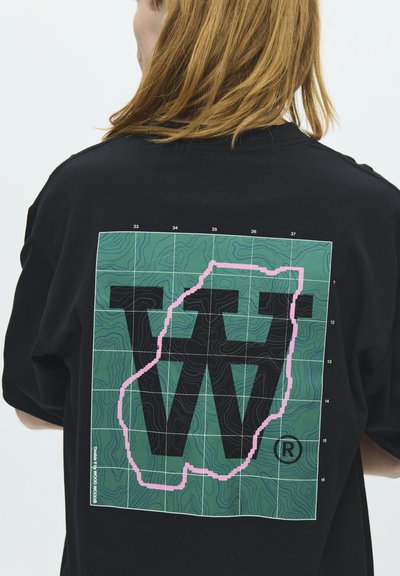 Personne aux cheveux châtain clair portant un t-shirt noir avec une carte en quadrillage vert et de grandes lettres superposées "V" et "W" bordées de rose.